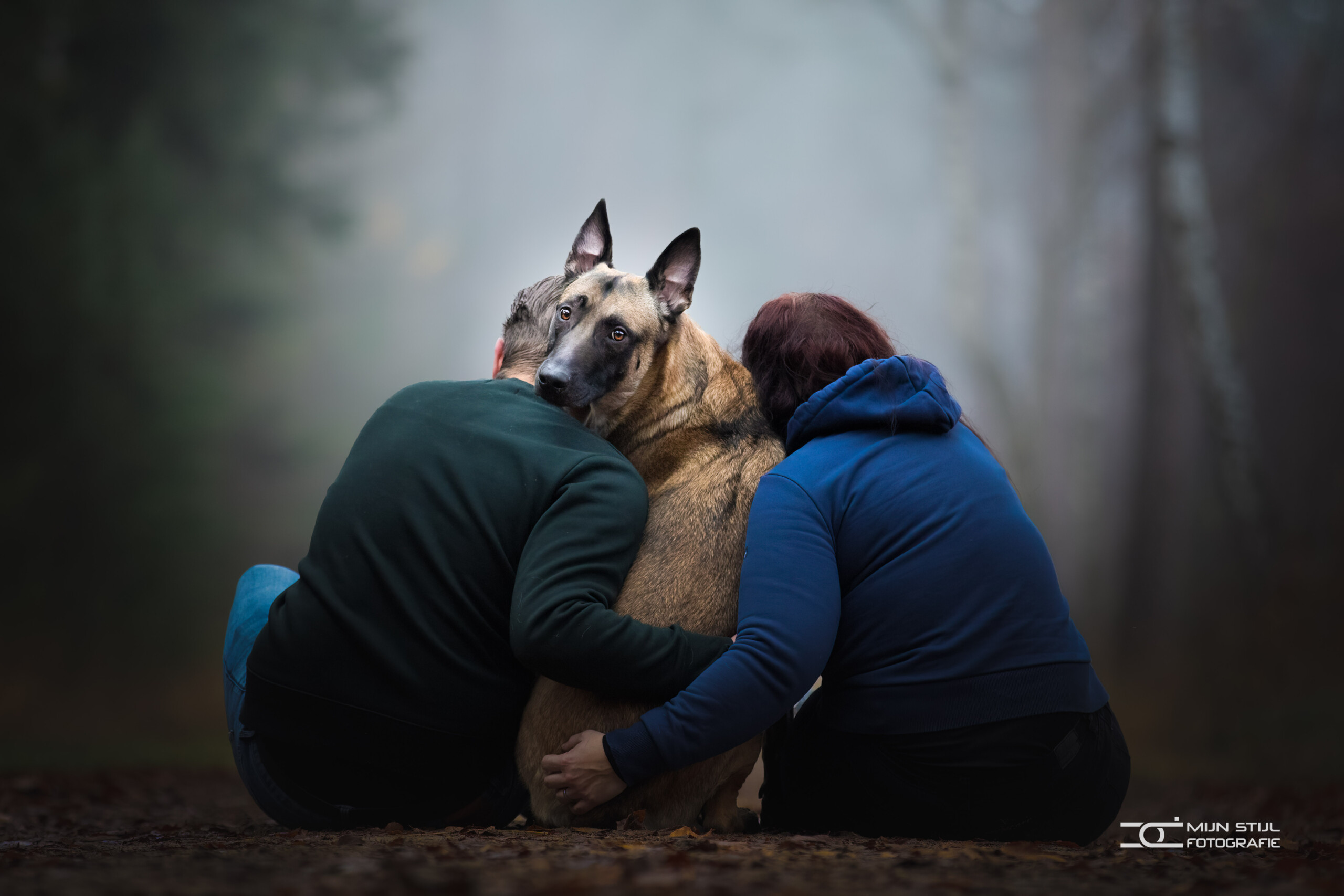 Een Mechelse herder wordt in het bos omarmd door twee baasjes.